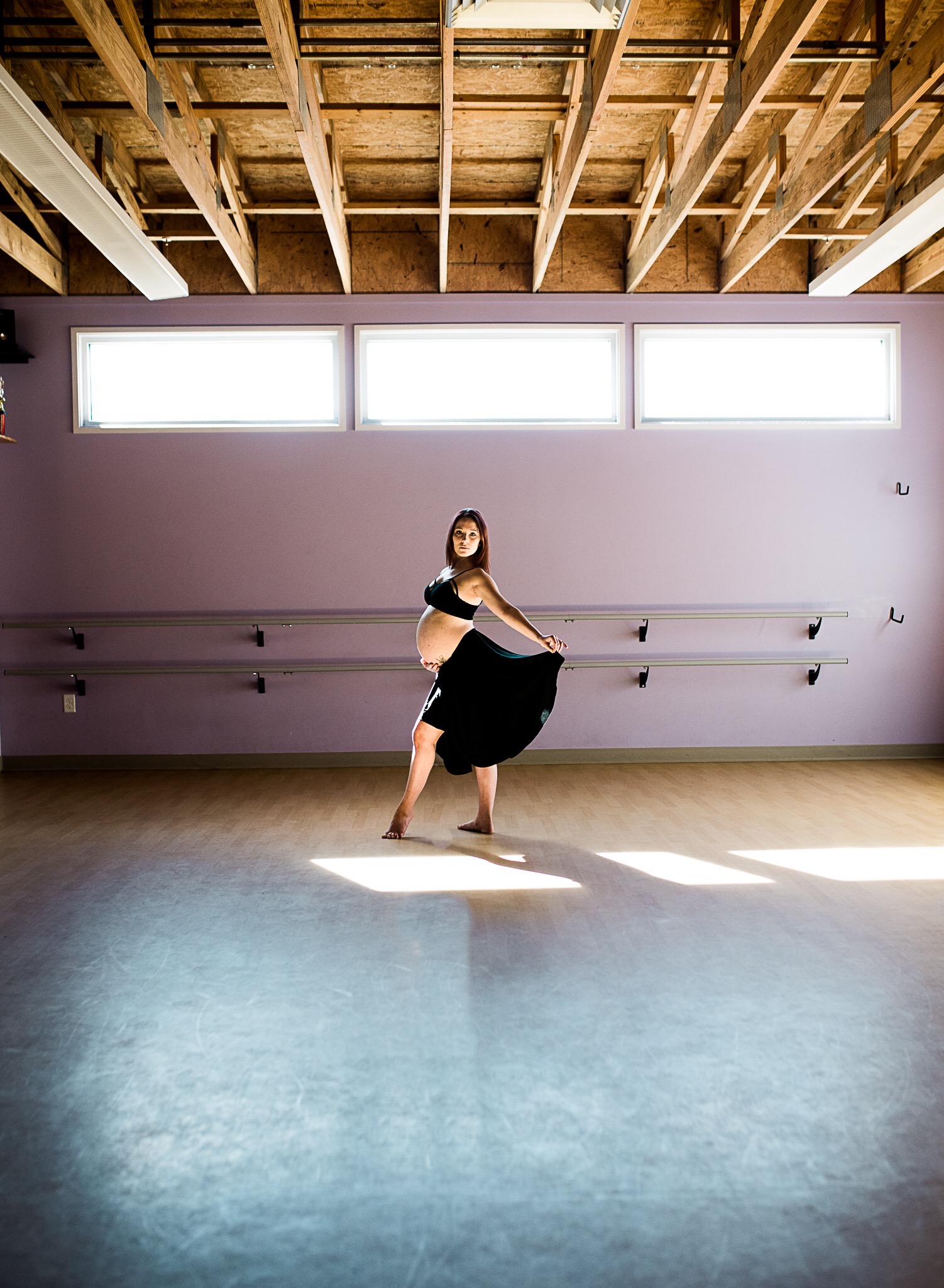 Becca at the barre in a black skirt, standing in a floor sunbeam in a bright, airy studio